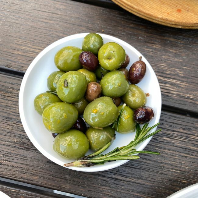A bowl of green and black olives with rosemary on a rustic wooden table outdoors.